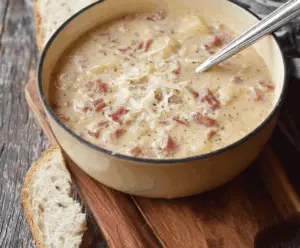 A steaming bowl of creamy Reuben soup garnished with shredded corned beef, Swiss cheese, sauerkraut, and fresh herbs, served in a white bowl on a wooden table.
